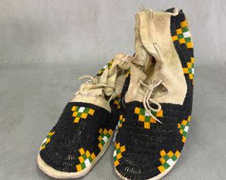 Pair of Native American beaded hide moccasins—likely 19th-century Lakota Sioux or Northern Plains origin—with sinew-sewn tanned hide and intricate beadwork of predominantly black vamp fields accented with distinctive geometric stepped or checkerboard beadwork in black, green, yellow, and white