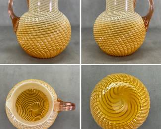 A charming vintage art glass pitcher, 8 inches high, featuring a twisted or reverse swirl optic pattern  warm amber-to-cream gradient and a distinct applied pink/peach-toned handle; possibly Buckeye Glass Co. or Fenton.