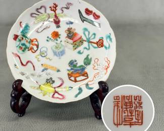 A Chinese export porcelain famille rose enameled dish, 5-3/4" diameter, decorated with various items including books, a ruyi staff, scroll, incense censer, fish bowl, peony, and a wrapped guqin (a seven-string zither.) The reverse decorated with flying bats in iron red, with six-character mark to the base.