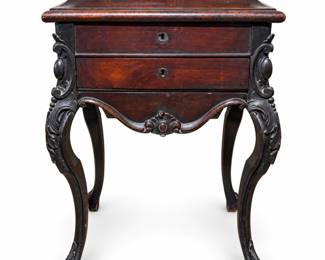 Antique nineteenth-century carved wood work or sewing table featuring a hinged lift-top opening to a fitted storage compartment, two locking front drawers, shaped apron and elaborately carved cabriole-style legs with scrolling foliate decoration. Rich dark wood grain and a warm aged finish give the piece considerable decorative character. Includes the pictured key. The upper compartment retains its wood interior and original-style hardware. Construction and overall form suggest a nineteenth-century work, sewing, dressing or personal storage table; exact maker, country of origin, wood species and date have not been confirmed. Displays substantial age-appropriate character including surface scratches, finish wear, abrasions, discoloration and interior wear as photographed. Please review all photographs carefully as they form an important part of the condition description.