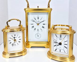 Collection of three decorative brass-tone desk and carriage-style clocks featuring classic white faces with Roman numeral hour markers. Includes a tall St. James London carriage-style clock with top handle and subsidiary dial, a Howard Miller quartz clock with decorative handle, and a Chass quartz desk clock with folding-style top handle. An attractive grouping with traditional styling suitable for an office, library, mantel, bedside table, or clock collection. All items are pre-owned and show light age and use consistent with display. Clocks have not been tested for timekeeping accuracy or long-term operation and are sold as-is. Please review photographs for condition and details.