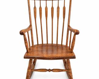 Antique Windsor-style wooden rocking chair featuring a tall spindle back with distinctive elongated paddle-form splats, broad curved crest rail, shaped wooden armrests, contoured solid wood seat, turned supports and stretcher, and traditional curved rocker runners. Finished in a warm medium-brown tone with attractive wood grain and an aged surface developed through years of use. The chair retains substantial character and displays visible age-related wear throughout, including scratches, scuffs, finish loss, marks and surface wear, particularly to the seat, arms and rocker runners. These imperfections contribute to its authentic antique appearance and have not been restored or concealed. Attractive country, farmhouse and Americana-style seating with a classic Windsor-inspired form. Please review all photographs carefully for construction, condition and signs of age.