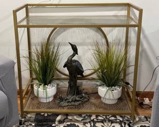 Two-tiered gold finished metal bar cart with clear glass shelves and caster wheels. Faux potted decor and heron lamp