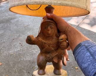 Pre-Columbian style Mexican folk art terracotta table lamp [index:1.1.10]. The ceramic base is modeled after a traditional Mesoamerican (often referred to as West Mexican, Colima, or stylized Mayan/Aztec) effigy figure [index:1.1.1], specifically depicting a chief or warrior holding a sacred bird under his arm 