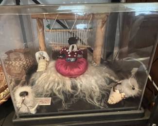 Native American style diorama featuring a doll at a weaving loom, a papoose, and small fur figures, housed in a clear acrylic display case.