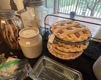 Vintage kitchenware collection featuring a Dazey Churn No. 40 glass butter churn, a stoneware crock, a ribbed glass dish, and decorative faux pies on a wire rack.