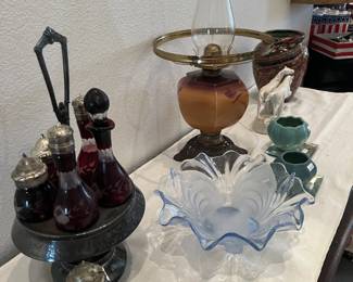 Assortment of vintage glassware and decor, including a ruby glass condiment set on a metal stand, a frosted blue glass ruffled bowl, and an antique-style oil lamp with a glass chimney.