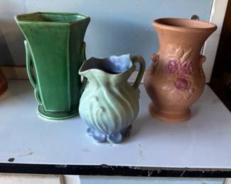 Three vintage art pottery vessels, including a green geometric McCoy Pottery vase, a small blue and green swirled Weller Pottery pitcher, and a terracotta-colored vase with embossed pink roses.