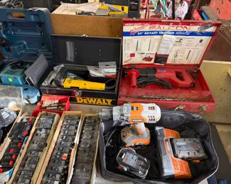 Assorted power tools including a Rigid cordless drill with charger, a DeWalt tool in a metal case, a Milwaukee rotary hammer in its original red metal box, and several trays of electrical circuit breakers.