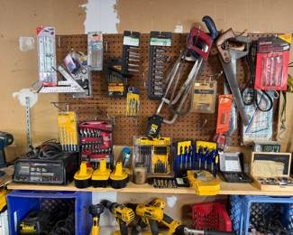 Assorted workshop tools and accessories, including a Dewalt Battery's & Battery charger, DeWalt power drills, hand saws, and various drill bit and socket sets organized on a pegboard and shelf.