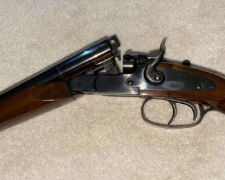 Close-up of a break-action double-barrel shotgun with a wooden stock, dual triggers, and an exposed hammer, showing the action in the open position.