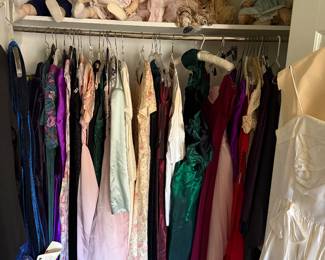 Closet of formal gowns with porcelain dolls and a teddy bear on the shelf above
