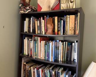 Dark bookcase of books topped with folded book art, a doll and a carved praying hands sculpture