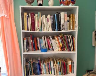 White bookcase filled with books, topped with a pheasant figurine, birdhouse, bud vase and Noah's Ark cookie jar