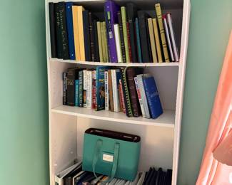 White bookcase with books, binders and photo albums, a vintage teal train case and carved decoys on top