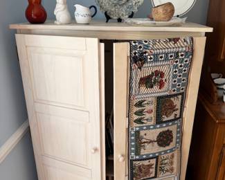 Light wood two door cupboard topped with an amber glass vase, rabbit figurine, creamer and decorative plates