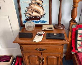 Nightstand with a turned wood lamp, clock radio and the framed ship embroidery