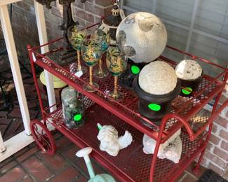 A red metal tea cart with hand painted wine glasses, several decorative items and cast iron standing cross.