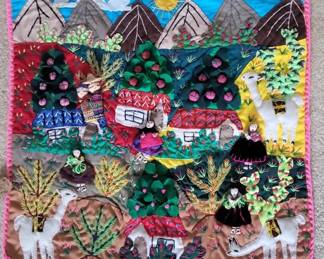 Colorful textile art featuring an embroidered or appliqué village landscape with houses, trees, and decorative elements in bright jewel tones and metallic accents. The piece has a decorative border and appears to be handmade or artisanal folk art.