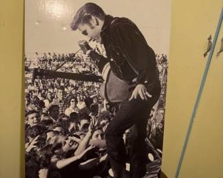 Black and white photograph of Elvis Presley performing on stage before a large crowd, mounted as a poster. The image captures the iconic performer in a characteristic pose during what appears to be a 1950s concert performance.