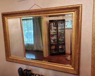 Large rectangular wall mirror with a gold-tone wooden frame. The mirror reflects a room interior and appears to be in good condition.