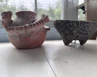 Two ancient artifacts: a terracotta vessel with a spout and decorative rim, and a dark volcanic stone mortar or grinding bowl. Both show significant age and wear consistent with pre-Columbian Mesoamerican origin.