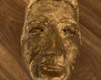 Hand-carved wooden mask with a naturalistic human face, finished in a golden or gilt tone. The piece shows detailed facial features and appears to be a decorative wall hanging or sculptural object.