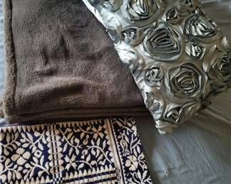 Lot of three textured throw blankets or pillows in neutral tones, including a brown knit piece, a patterned black and white geometric design, and a quilted metallic silver piece.