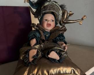 Porcelain head doll dressed in a black and gold jester or harlequin costume with a decorative hat. The doll has painted facial features and appears to be seated on a gold-colored cushion.