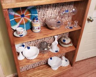 Open wooden bookshelf with three shelves displaying a mix of decorative items including ceramic pieces, glassware, and a blue and orange decorative plate. The shelves are lined with cork or similar material backing.
