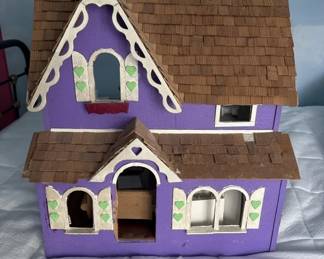 Two-story purple wooden dollhouse with cream and brown trim, featuring multiple rooms with windows and doorways. The structure appears handmade or custom-built with a peaked roof design.