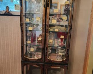Two-door wooden display cabinet with glass fronts and shelving, containing various decorative items and collectibles. The cabinet features a dark wood frame with lower solid wood panels and upper glass sections for displaying items.