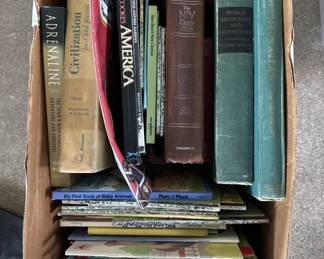 A cardboard box containing approximately 10-12 books of various sizes and subjects, including hardcover and paperback volumes. Spines show titles related to history, literature, and other topics.