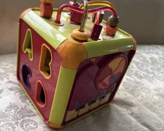 Colorful tin cube toy with red, yellow, and green panels featuring cutout shapes and small wooden pegs or handles on top. Appears to be a mid-20th century children's activity toy.