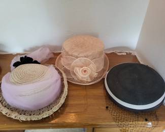 Three vintage women's hats in various styles and colors, including a lavender hat with netting, a tan/beige hat with a decorative bow, and a black hat with white trim. All appear to be mid-20th century dress hats.