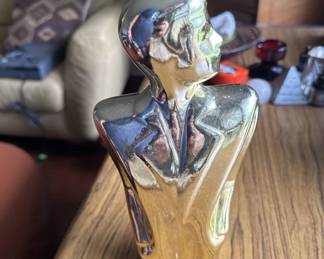 Hand-carved wooden figure with flowing abstract form, featuring polished metal inlays or overlays in silver and dark tones. The piece appears to be a modernist or contemporary art sculpture mounted on a wooden base.