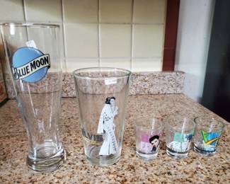 Blue Moon branded pilsner glass alongside four smaller decorative drinking glasses with various designs. All appear to be in good condition.