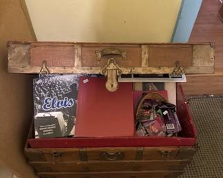 Early-to-mid 20th century steamer trunk with rounded top and metal hardware, containing various items including what appears to be vintage clothing and accessories. The trunk shows wear consistent with age and use.