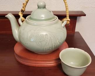 Pale green celadon teapot with relief-carved floral design, domed lid, and woven rattan handle, accompanied by a matching small cup. The set appears to be Asian ceramic work, likely Chinese or Vietnamese in origin.