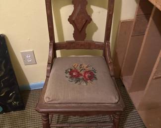 Wooden chair with a decorative needlepoint seat featuring a red floral design. The chair has a tall back with carved details and appears to be in used condition.