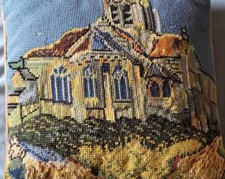 A needlepoint or tapestry piece featuring a detailed architectural scene of a church or cathedral building with yellow and blue tones. The work appears to be hand-stitched on canvas with traditional needlework techniques.