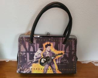 Vintage-style handbag featuring Elvis Presley imagery with guitar and performance graphics on a purple background. Black handles and structured frame design.
