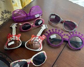 Lot of approximately 8 novelty and costume sunglasses in various styles and colors, including guitar-shaped frames, oversized round frames, and decorative designs in purple, pink, and white. Most appear to be unworn or lightly used.