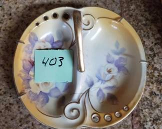 Round porcelain plate featuring hand-painted blue and purple floral motifs with ornamental scroll handles on either side. The piece has a cream-colored background with gilt accents.