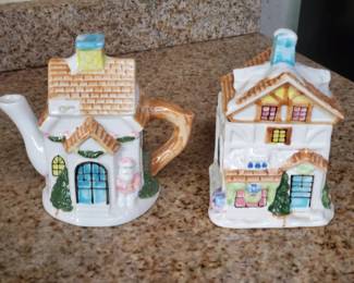 Two-piece ceramic tea set featuring a teapot shaped like a cottage with a handle and spout, paired with a matching house-shaped creamer. Both pieces are decorated with painted details including windows, doors, and landscaping elements.