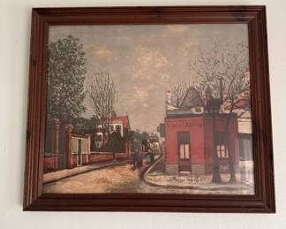 Framed artwork depicting a vintage street scene with period buildings, trees, and figures. The piece is housed in a dark wood frame and appears to be a print or painting on canvas.