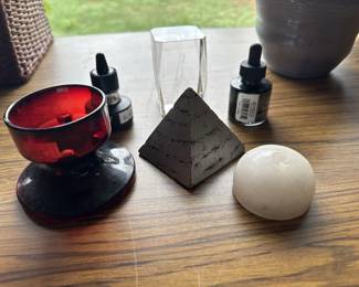 Collection of metaphysical items including a black pyramid crystal, white sphere, red glass bowl with stand, clear quartz point, and two dark glass bottles with droppers, typically used for essential oils or tinctures in alternative wellness practices.