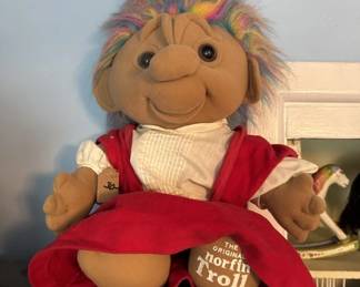 A vintage troll doll with orange yarn hair, painted facial features, and a red dress with white collar. The doll appears to be from the 1960s-1970s era based on its style and construction.