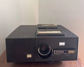 Vintage Keystone 35mm slide projector with black metal housing and manual focus lens. Appears to be in working condition with minor wear consistent with age.