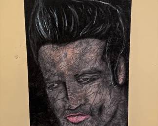 Fabric or textile art depicting Elvis Presley with his iconic black pompadour hairstyle and pink lips. The piece appears to be printed or embroidered on dark fabric.